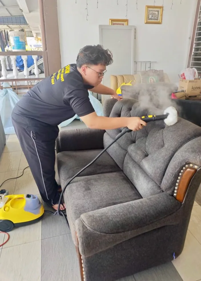 Pekerja mencuci sofa kelabu dengan mesin wap di ruang tamu rumah pelanggan